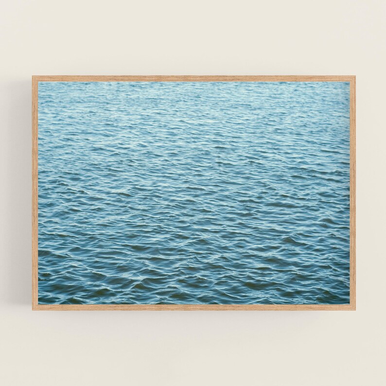 May include: A framed print featuring a close-up view of rippling blue water. The water's surface is textured with small waves, creating a sense of depth and movement. The wooden frame adds a natural touch to the artwork.