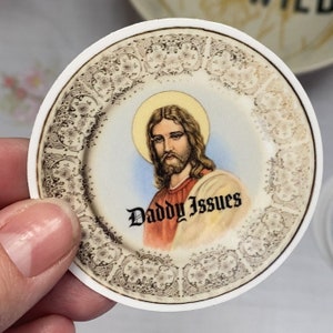 May include: A round sticker featuring an image of Jesus Christ with a golden halo, set against a decorative plate design. The text "Daddy Issues" is written in a gothic font below the image. The sticker is held by a hand.