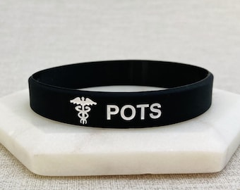 Puede incluir: Pulsera de silicona negra con un símbolo médico blanco y el texto "POTS" en blanco.