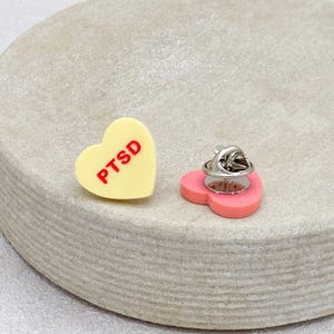 May include: Two heart-shaped pins. One is yellow with the red letters "PTSD". The other is pink with a silver pin back. Both pins are on a light gray, textured surface.