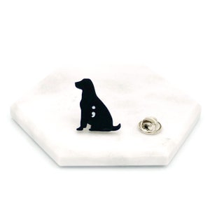 May include: Black enamel pin of a dog sitting with a semicolon on its side. The pin is on a white marble surface with a silver pin back.