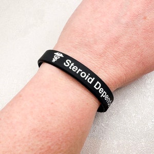 Peut inclure: Bracelet en silicone noir avec du texte blanc qui dit "Steroid Dependent" et un symbole m&eacute;dical.