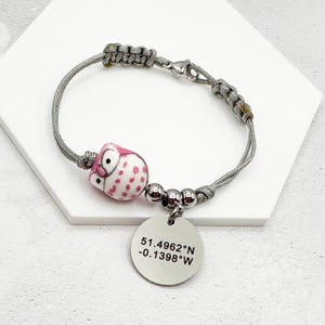 May include: A gray cord bracelet with a pink owl charm and a silver charm with the coordinates 51.4962°N -0.1398°W.