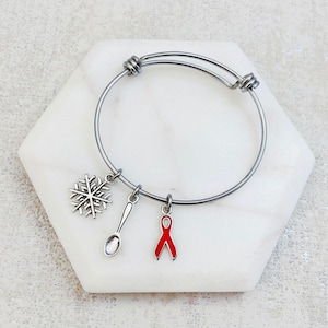 May include: A silver bangle bracelet with three charms: a snowflake, a spoon, and a red awareness ribbon.