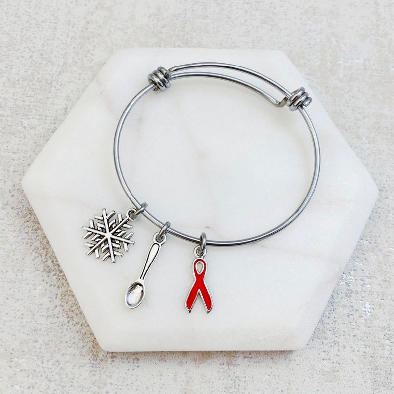 May include: A silver bangle bracelet with three charms: a snowflake, a spoon, and a red awareness ribbon.