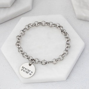 Könnte beinhalten: Ein silberfarbenes Armband mit Kettenglied-Design und einem runden Anhänger. Der Anhänger ist mit den Koordinaten 52°41'22" N 2°01'55" W graviert. Das Armband liegt auf einer weißen sechseckigen Oberfläche.