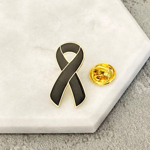 Könnte beinhalten: Schwarze Schleifen-Anstecknadel mit goldener Rückseite. Die Schleife ist ein Symbol für Trauer und Erinnerung.