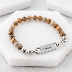 Può includere: Un braccialetto di perline di pietra marroni e beige con una chiusura argentata e un'etichetta argentata incisa con il nome "Tanya".