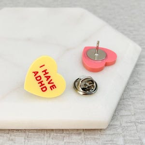 Pode incluir: Um conjunto de três alfinetes de esmalte em forma de coração. Um alfinete amarelo com o texto "I HAVE ADHD" em vermelho, um alfinete rosa com um fecho de alfinete prateado e um fecho de alfinete prateado.