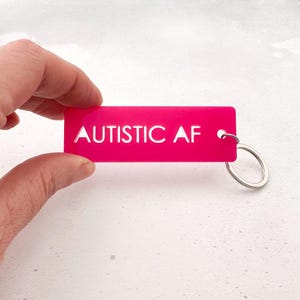 May include: A bright pink rectangular keychain with the words "AUTISTIC AF" cut out in white. The keychain has a silver key ring attached to the right side. The keychain is held by a hand.