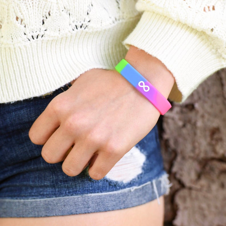 K&ouml;nnte beinhalten: Ein regenbogenfarbenes Silikonarmband mit einem wei&szlig;en Unendlichkeitszeichen. Das Armband wird am Handgelenk getragen.