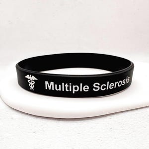 Può includere: Un braccialetto in silicone nero con il testo bianco Multiple Sclerosis e un simbolo medico caduceo bianco stampato sul davanti. Il braccialetto è circolare e ha una finitura opaca liscia, con scritte chiare e leggibili e un'icona distinta.