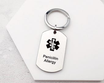 May include: A silver-colored metal keychain with a dog tag-shaped pendant. The pendant features a black medical alert symbol and the words "Penicillin Allergy" in black text. The keychain has a circular ring for attaching keys.