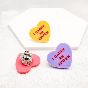 May include: Heart-shaped enamel pins in yellow and purple, with the text "I CARRY AN EPIPEN" in red. One pin is attached to a pink heart-shaped base with a silver pin back. The pins are on a white surface.