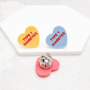 Può includere: Tre spille a forma di cuore in smalto. Una è gialla, una blu e una rosa. Ogni spilla riporta la scritta "TYPE 1 DIABETES" in rosso. Le spille hanno una chiusura in metallo sul retro.