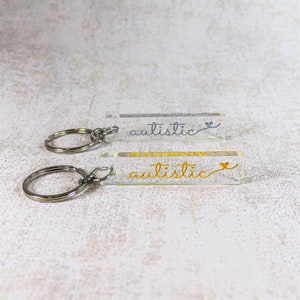May include: Two clear acrylic keychain charms with silver key rings. The charms are rectangular and have the word "autistic" written in cursive on them. One charm has the word in silver glitter and the other in gold glitter.