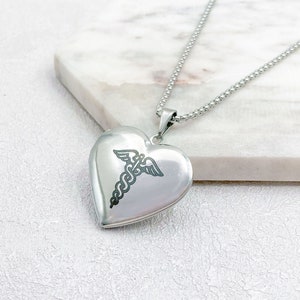 May include: A silver heart-shaped locket necklace with a medical caduceus symbol engraved on the front. The pendant hangs from a delicate chain. The locket is displayed on a white marble surface.