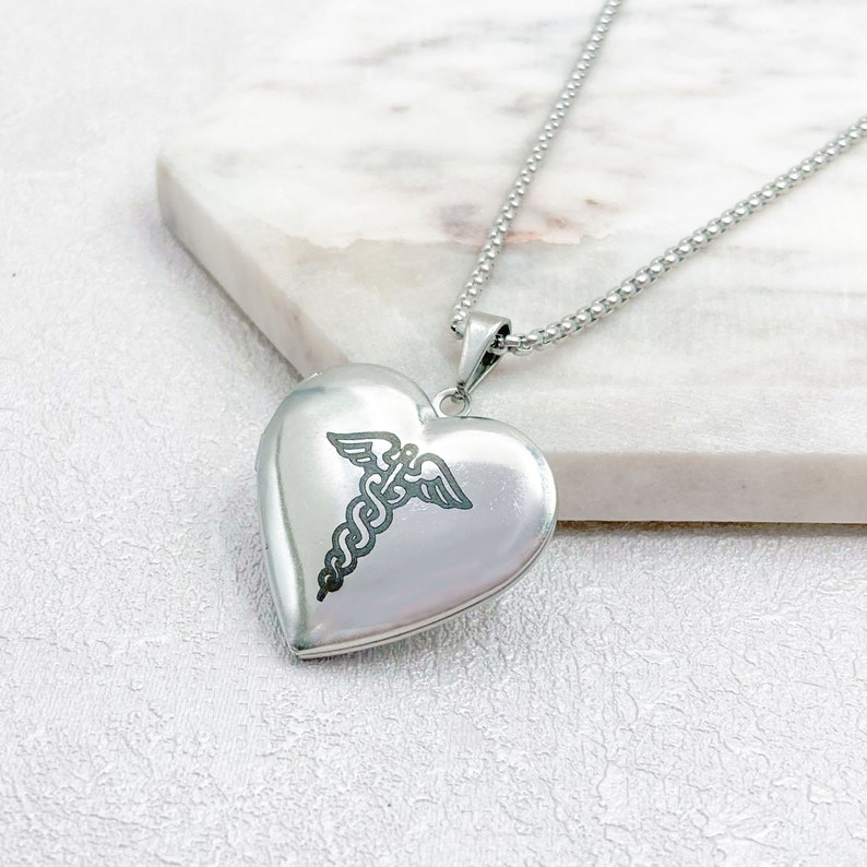 May include: A silver heart-shaped locket necklace with a medical caduceus symbol engraved on the front. The pendant hangs from a delicate chain. The locket is displayed on a white marble surface.