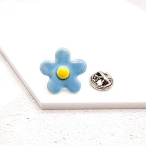 May include: A light blue flower-shaped enamel pin with a yellow centre. The pin has a silver-coloured backing for attachment. The flower design is a cheerful accessory.