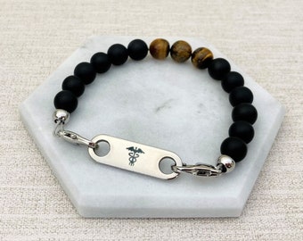 Puede incluir: Pulsera de cuentas negras y ojo de tigre con una placa de identificación médica de acero inoxidable grabada con el símbolo del caduceo.
