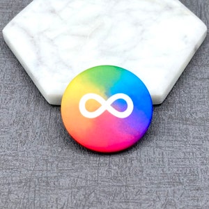 K&ouml;nnte beinhalten: Ein runder Knopf mit einem Regenbogen-Farbverlauf als Hintergrund und einem wei&szlig;en Unendlichkeitszeichen in der Mitte.