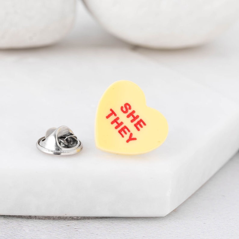 Close-up of She/They pronoun pin with dual pronoun design on acrylic heart