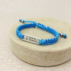 May include: A blue braided bracelet with a silver tag that reads "2° 26'33"W 52° 56'40"N".
