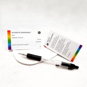 May include: Two white cards with text, one reading "IN CASE OF EMERGENCY" with a rainbow stripe, and the other saying "HELLO, I AM AUTISTIC." A clear pen with a black grip rests on a white, round surface.