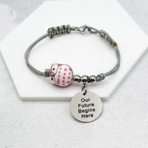 May include: Gray cord bracelet with a pink and white owl charm and a silver charm that says "Our Future Begins Here".