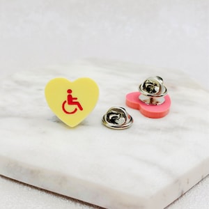 Peut inclure: Une épingle en forme de cœur jaune avec un symbole d'accessibilité en fauteuil roulant rouge. L'épingle est fixée à un dos d'épingle argenté.