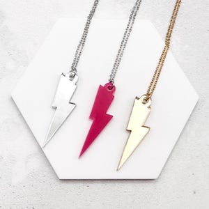 Könnte beinhalten: Drei Halsketten mit Blitzanhängern. Eine ist silberfarben, eine pinkfarben und die dritte goldfarben. Jeder Anhänger hängt an einer filigranen Kette. Die Halsketten werden auf einer weißen, geometrischen Oberfläche präsentiert.