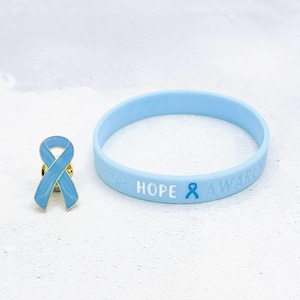 Puede incluir: Un broche de cinta azul claro y una pulsera de goma a juego. El broche tiene forma de cinta con un contorno dorado. La pulsera tiene las palabras "HOPE AWARENESS" y un gráfico de cinta. Ambos artículos están sobre una superficie blanca.
