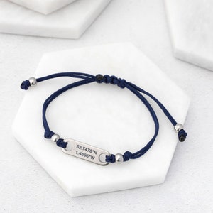 May include: A navy blue adjustable bracelet with a silver-colored rectangular charm. The charm is engraved with the coordinates 52.7478°N 1.4696°W. The bracelet features silver beads and a sliding knot closure.