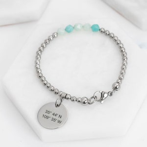 Könnte beinhalten: Ein silbernes Armband mit Perlen und einem Anhänger, auf dem die Koordinaten 35° 44' N 109° 35' W eingraviert sind. Das Armband hat hellblaue und weiße Perlen.