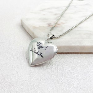 May include: A silver heart-shaped locket pendant on a chain. The locket has the words "Type 1 Diabetes" engraved on the front. The necklace is displayed on a white marble surface.