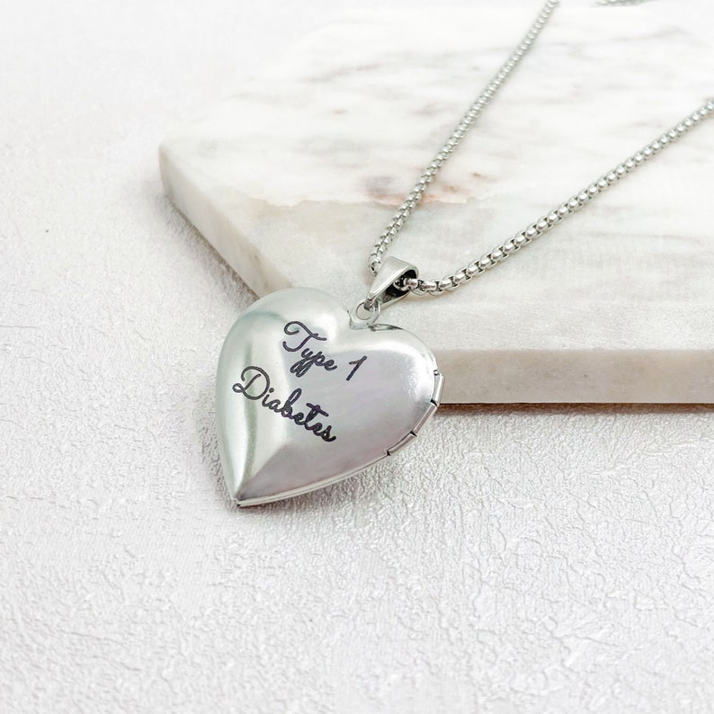 May include: A silver heart-shaped locket pendant on a chain. The locket has the words "Type 1 Diabetes" engraved on the front. The necklace is displayed on a white marble surface.