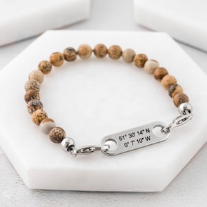 Op de afbeelding: Een bruin en beige stenen kralen armband met een zilveren tag gegraveerd met de coördinaten "51° 30' 14" N 0° 7' 10" W".