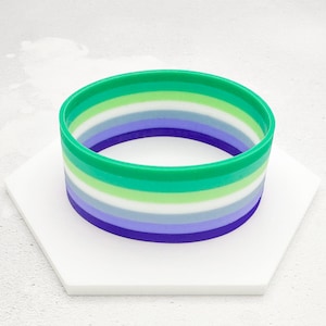May include: A green, white and purple striped rubber bracelet. The bracelet is a symbol of the genderqueer pride flag.
