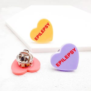 May include: Three heart-shaped enamel pins. One is yellow with red text that reads "EPILEPSY". Another is purple with the same text. The third pin is pink with a silver pin back.