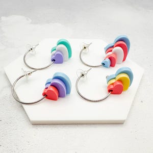 Puede incluir: Cuatro pares de pendientes de aro con colgantes en forma de corazón de colores. Cada pendiente presenta un aro plateado con un grupo de corazones en capas en tonos de rojo, amarillo, azul, morado y verde. Los pendientes se exhiben sobre una superficie hexagonal blanca.