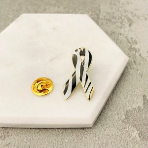 Pode incluir: Um alfinete dourado com um design de fita listrada em preto e branco em forma de zebra. O alfinete está sobre um fundo branco.