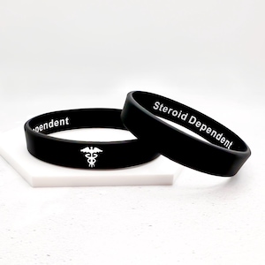 Pode incluir: Duas pulseiras de silicone preto. Uma pulseira diz "Steroid Dependent" em texto branco. A outra pulseira tem a palavra "dependent" e um símbolo de caduceu branco. As pulseiras são exibidas em uma superfície branca.