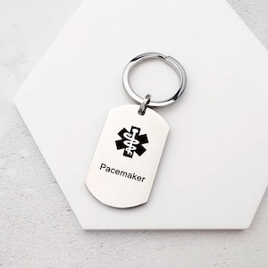 Könnte beinhalten: Ein silberfarbener Schlüsselanhänger mit einem schwarzen medizinischen Symbol und dem Wort "Pacemaker" darunter. Der Schlüsselanhänger hat einen runden Ring zum Befestigen an Schlüsseln.