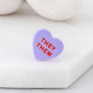Close-up of They/Them pronoun pin showing engraved lettering on acrylic heart badge