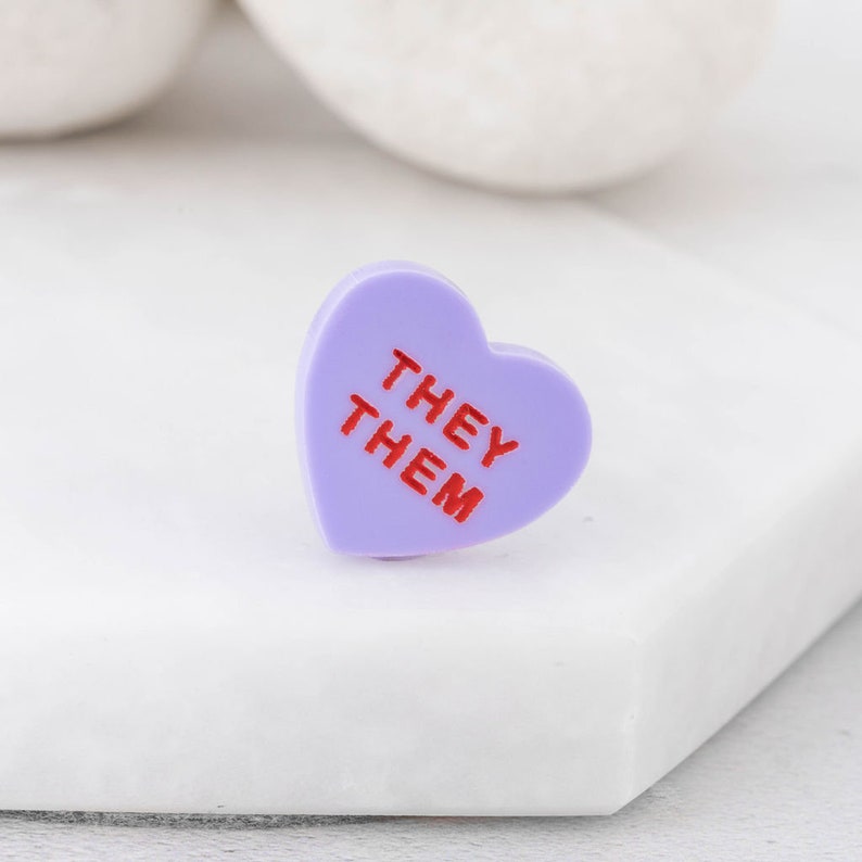 Close-up of They/Them pronoun pin showing engraved lettering on acrylic heart badge