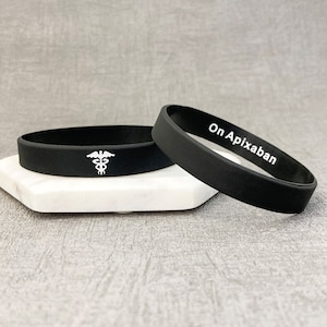 May include: Two black silicone bracelets. One bracelet has a white medical symbol, the other has the text "On Apixaban" in white.