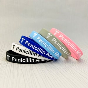 Puede incluir: Un conjunto de seis pulseras de silicona en negro, blanco, azul, azul claro, gris y rosa. Cada pulsera tiene un símbolo médico blanco y el texto "Penicillin Allergy".