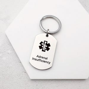 Może przedstawiać: Srebrny brelok do kluczy w kształcie nieśmiertelnika z czarnym symbolem medycznym i napisem "Adrenal Insufficiency". Brelok posiada srebrny pierścień do mocowania kluczy.