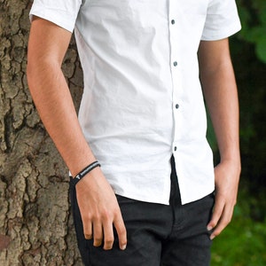 Puede incluir: Una persona con una camisa blanca abotonada con botones negros y pantalones negros. Lleva una pulsera negra con el texto "Immunosuppressant" en ella.