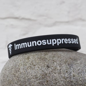 Puede incluir: Pulsera de silicona negra con el texto "Inmunodeprimido" y un s&iacute;mbolo m&eacute;dico blanco.